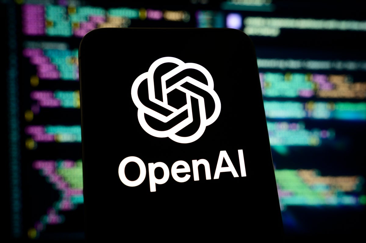 OpenAI logo and name on a smartphone against blurred, multicolored computer code on monitors.