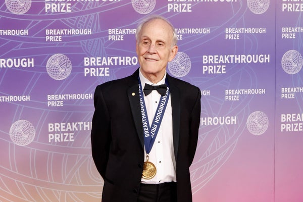 The physicist David J. Gross stands in formal wear in front of a wall emblazoned with Breakthrough Prize logos, and wears a Breakthrough Prize medal around his neck.