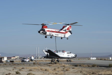 A large helicopter hovers over a tethered Dream Chaser space plane on an airport runway.