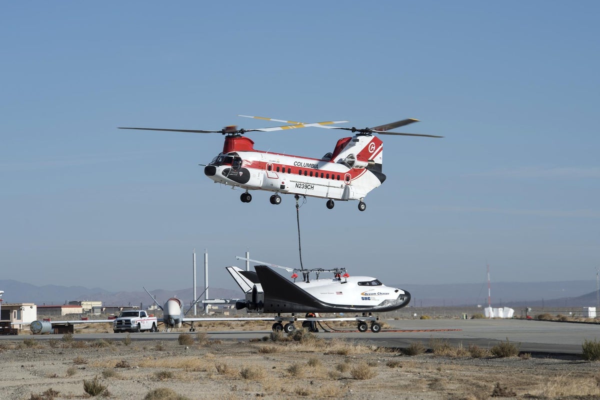 A large helicopter hovers over a tethered Dream Chaser space plane on an airport runway.