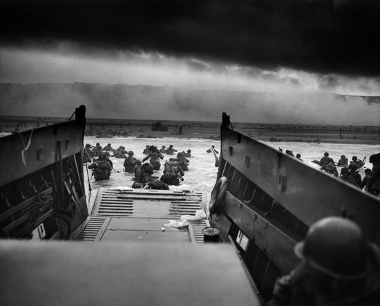 A black-and-white photo of soldiers leaving a troop transport to wade into the ocean toward the beach where smoke rises