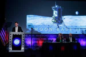 Isaacman stands behind a podium onstage to the left of a screen with a moon base graphic.