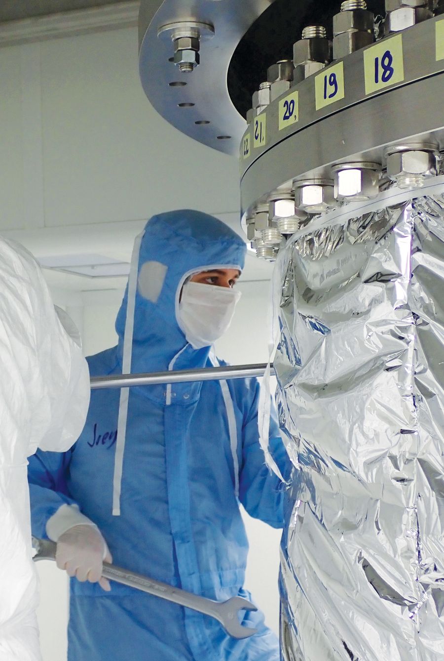 The author, wearing a blue full-body protective suit and white dusk mask, stands beside a large instrument sheathed in shining metal foil holding a large metal wrench. Provided by Elena Aprile
