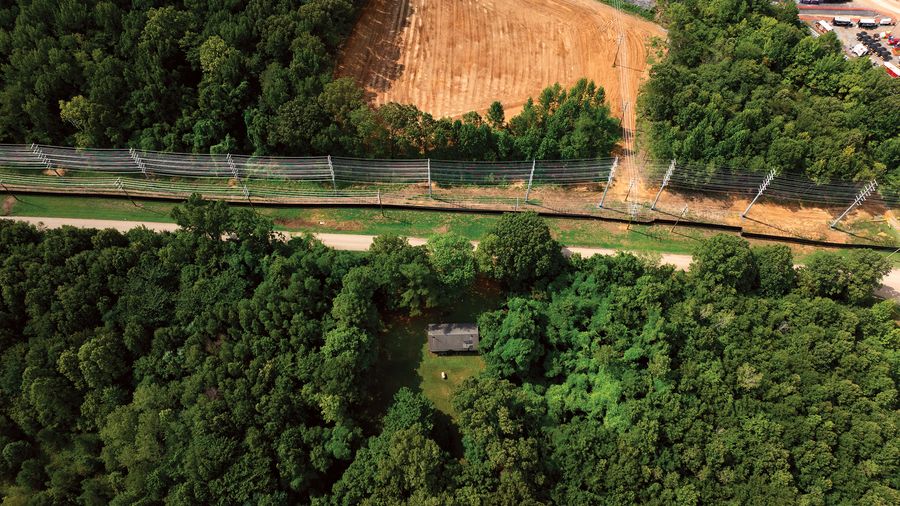From above, a small house sits in a clearing among dense trees, with a road, power lines and an industrial site just beyond.