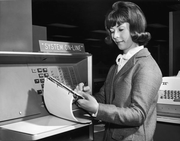 A woman with a clipboard stands beside a mainframe computer under a sign reading SYSTEM ON-LINE.