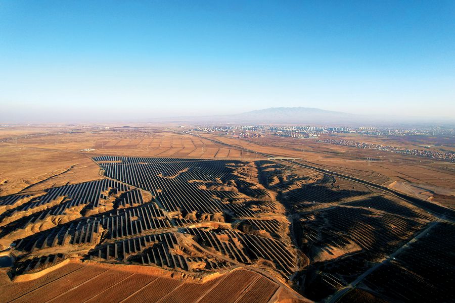 An oblique aerial view of solar panels on undulating brown hills with buildings and mountains shrouded in haze in the background. Image provided by VCG/Getty Images.