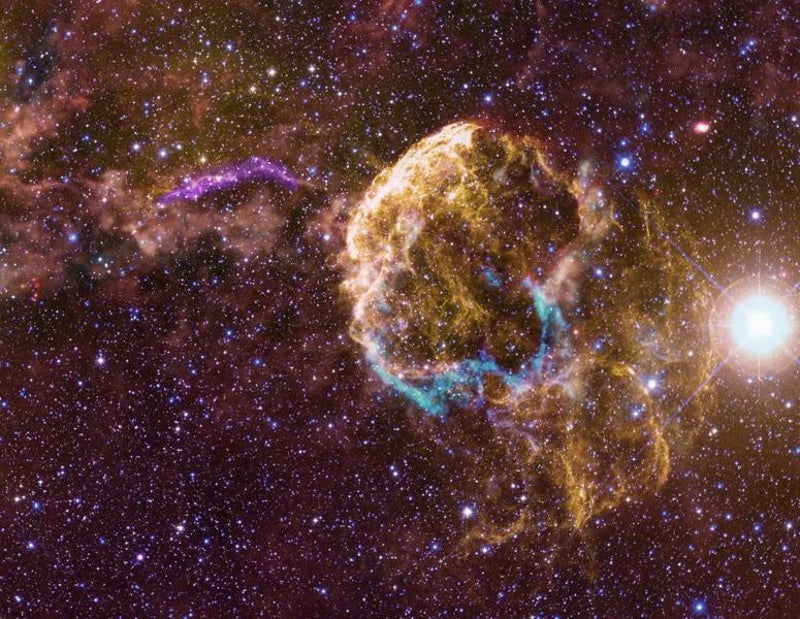 On a mottled reddish background speckled with stars, yellow threads of gas at center right trace out a dome-shaped structure from which curving tendrils extend toward the lower right. Blue and red filaments trace a ring around the dome&rsquo;s base. A violet arc stretches right to left just above the center of the image, and irregular orange gas clouds become thicker toward the upper left.