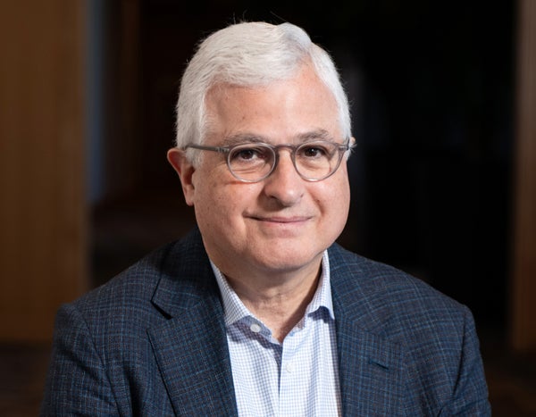 Close up of a man with white hair and glasses in a navy suit.