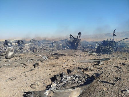 Burned-out wreckage of an aircraft scattered across a barren desert landscape, with twisted metal debris, charred components, and light smoke rising in the distance under a clear blue sky.