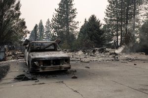 A burned out truck seen from the front, with debris and evergreen trees behind it against a smoky gray sky
