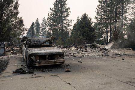A burned out truck seen from the front, with debris and evergreen trees behind it against a smoky gray sky