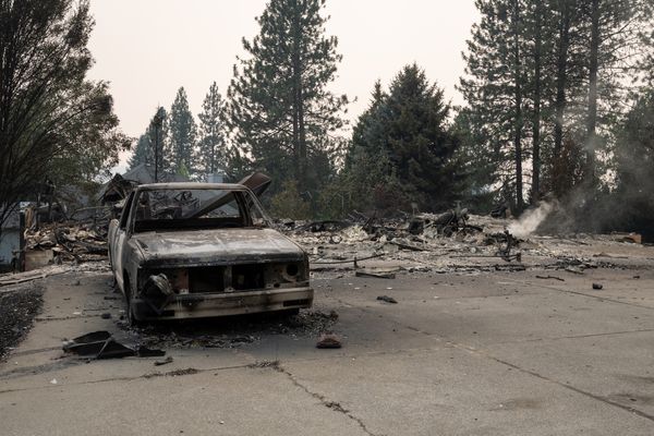 A burned out truck seen from the front, with debris and evergreen trees behind it against a smoky gray sky