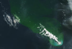 A plume of white smoke and steam streams from the bottom right corner of the image toward the top left. The water in the top and right of the image is a bright green, the bottom left dark blue. A small circle of brownish gray surround the area where the smoke plume emerges.