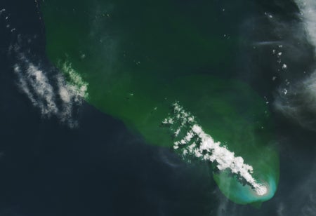 A plume of white smoke and steam streams from the bottom right corner of the image toward the top left. The water in the top and right of the image is a bright green, the bottom left dark blue. A small circle of brownish gray surround the area where the smoke plume emerges.