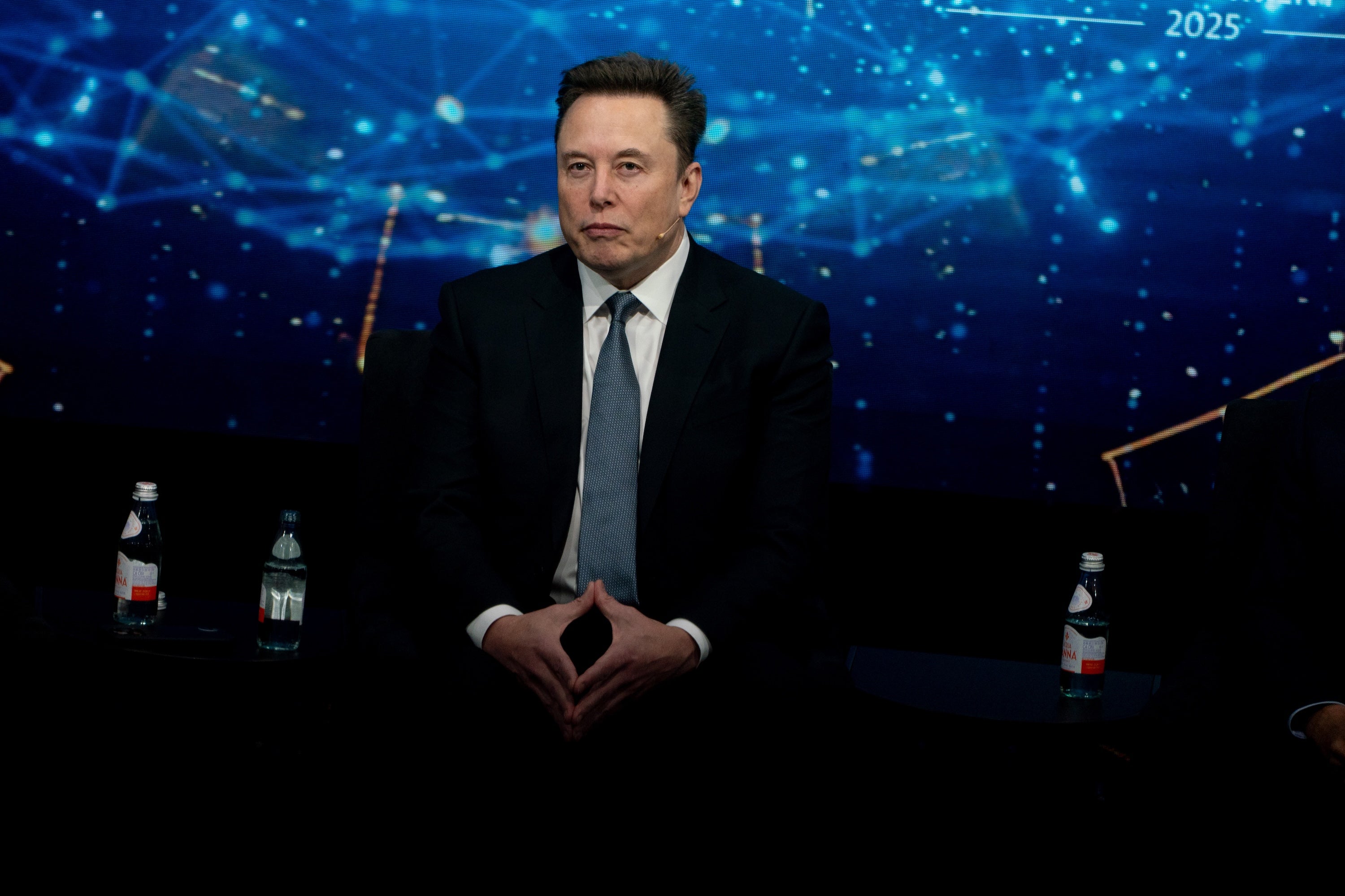 Clad in business attire, Elon Musk sits in front of a digital screen displaying abstract art.