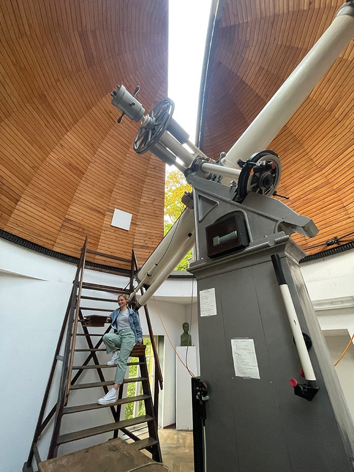 Astrophysicist Sydney Erickson stands on a wooden ladder next to a historical telescope inside a small room with a domed ceiling in Munich.