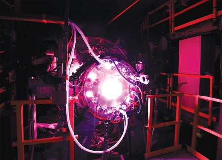 A cylindrical fusion prototype glows white and pink inside a dark test bay, surrounded by cables, hoses and metal supports.