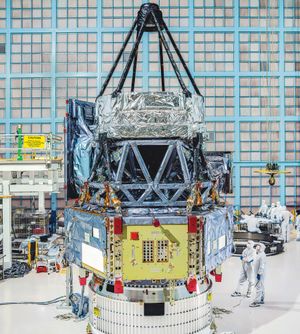 The foil-covered Roman telescope stands in a clean room surrounded by workers in bunny suits preparing it for launch. Image provided by NASA/Chris Gunn.