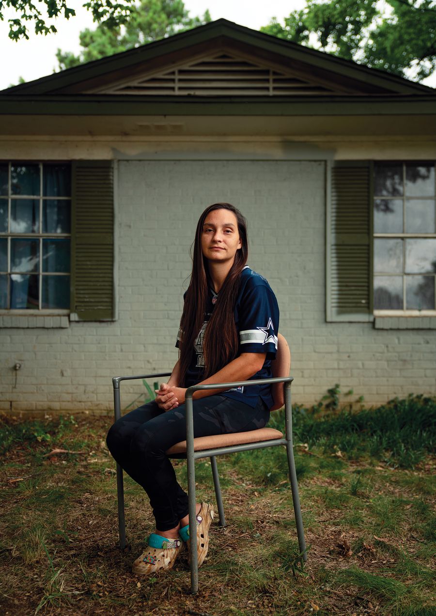 A woman in a dark football jersey sits in a folding chair beside a one-story brick house.