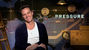 Andrew Scott cocks his head and smiles. He is sitting in front of an old-timey sea map and is wearing a white shirt, a dark blue cardigan and two thin gold necklaces. The word "PRESSURE" is next to him in all capital letters.