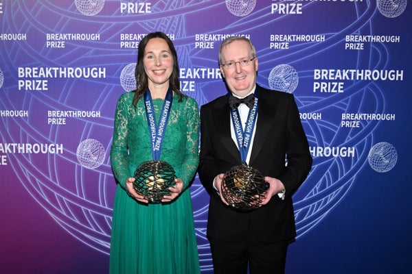 Rosa Rademakers and Bryan Traynor stand in formal dress in front of a Breakthrough Prize backdrop. They are wearing medals and holding an award.