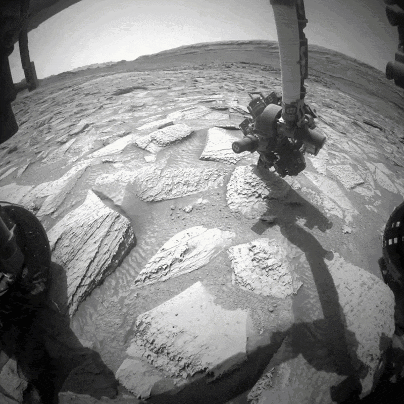 A black-and-white GIF of the Curiosity mars rover shaking off a large rock stuck to its robotic arm.