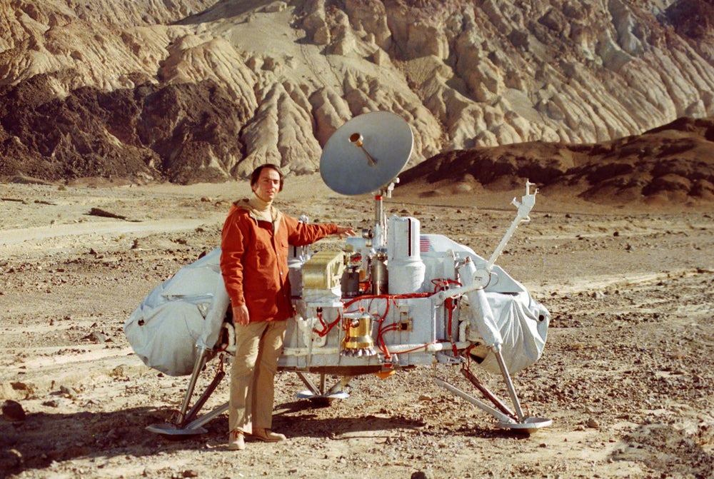 A man stands in front of a robotic interplanetary lander in a mountainous desert.