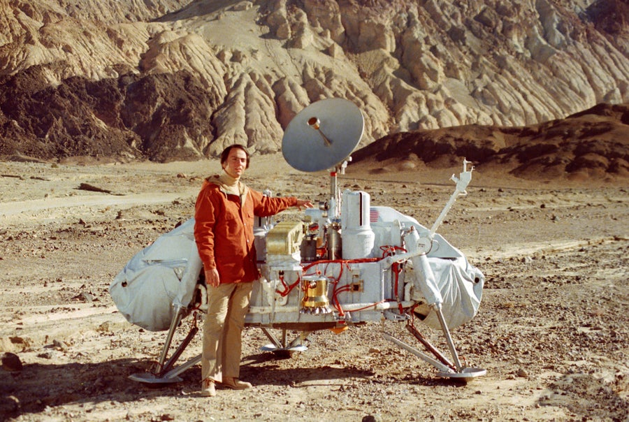 A man stands in front of a robotic interplanetary lander in a mountainous desert.