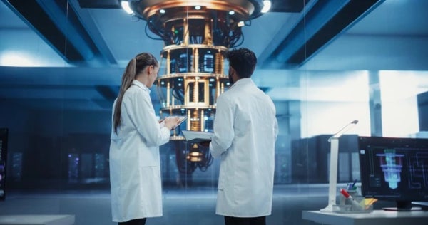 A woman and a man, with their backs towards the viewer, wearing lab coats and standing in front of a quantum computer.