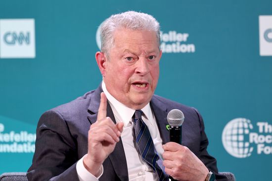 An older white man sits holding a microphone in one hand and pointing upward with another in front of a teal background
