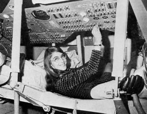 A woman in glasses sitting inside a spacecraft in black and white.