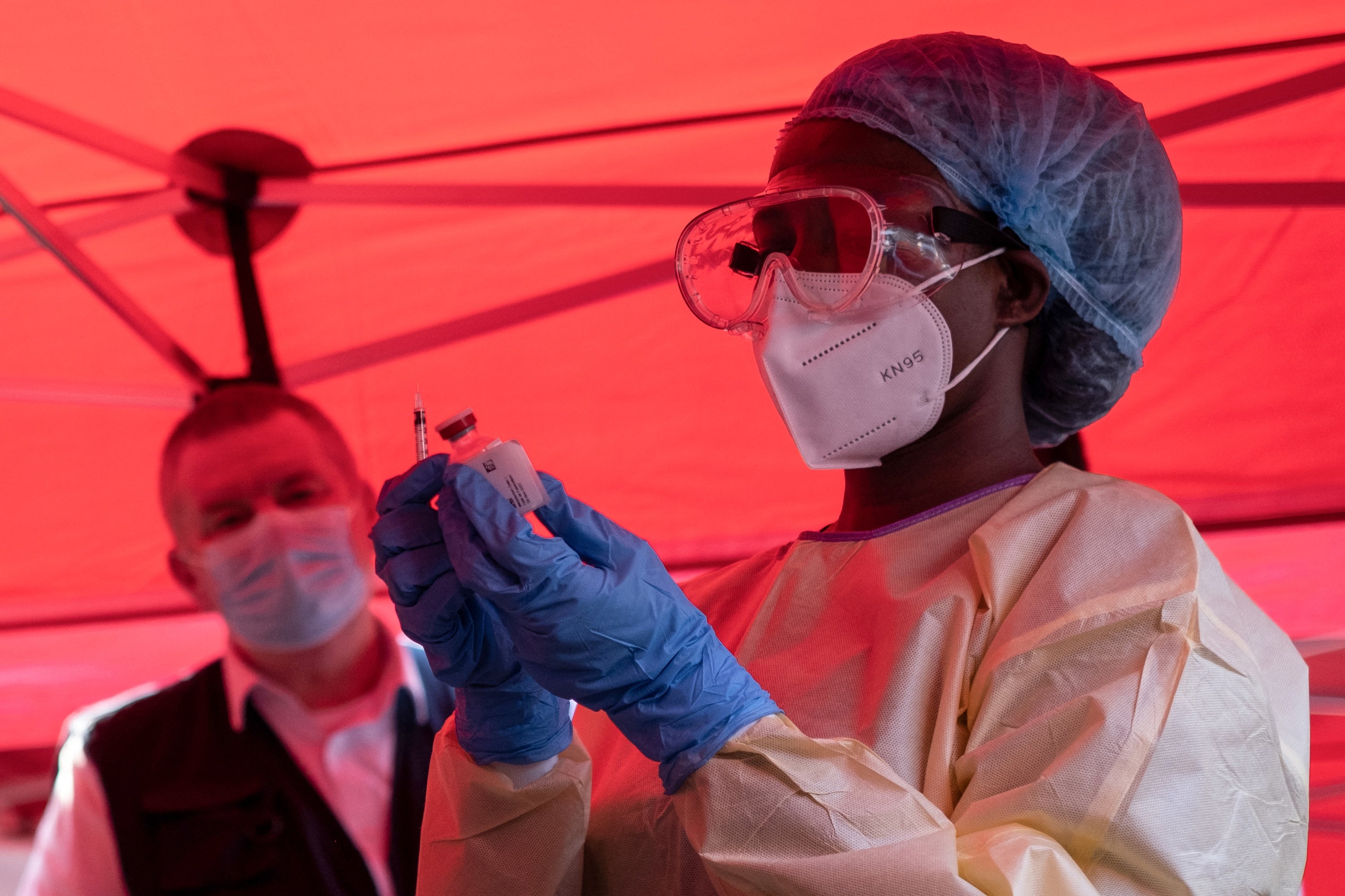 A nurse wearing protective medical clothing prepares a syringe by drawing a dose from a vaccine vial inside a red tent.