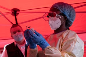 A nurse wearing protective medical clothing prepares a syringe by drawing a dose from a vaccine vial inside a red tent.