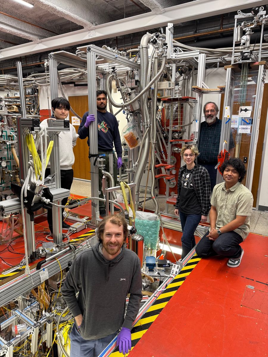 Six physicists pose around a series of wires and metal machinery in a physics lab.