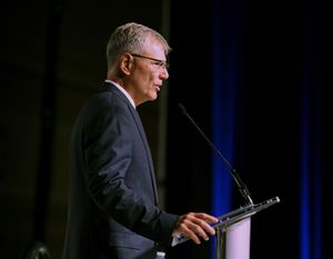 A middle-aged man wearing eyeglasses and a suit is seen in profile speaking at a lectern in a darkened auditorium.