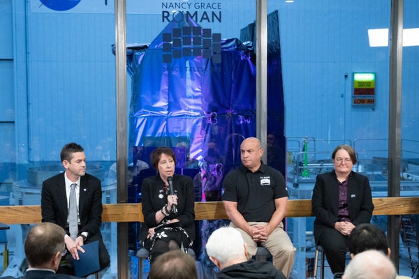 Four people sitting on tall chairs with a large spacecraft visible in the glass wall window behind them.