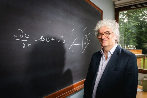 A man with glasses and curly white hair stands to the right of a blackboard with an equation written on it.