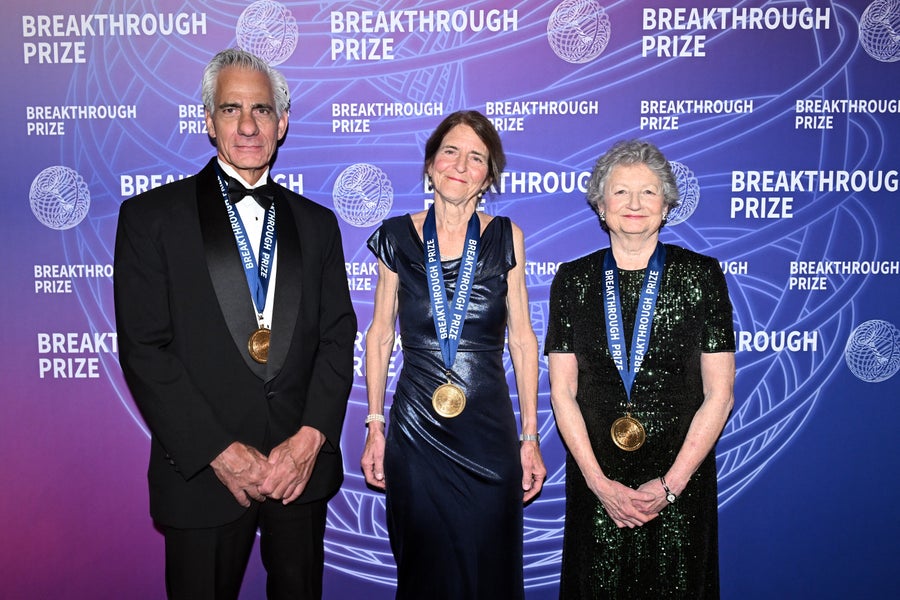 two women and one man in formal attire wearing gold medals. they stand in front of a press junket, the background reading breakthrough prize