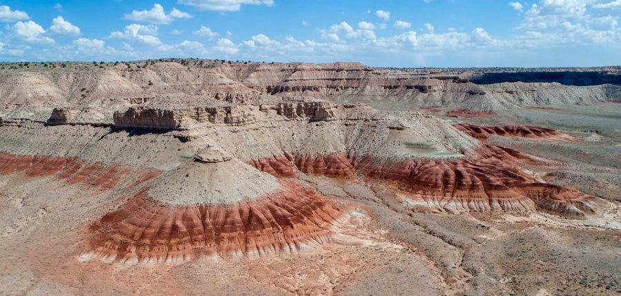 An aerial view of a mesa with tan layers atop a ring of red with a blue sky with fluffy white clouds in the background
