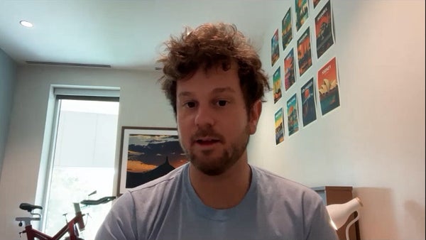 A man with light skin and auburn hair wearing a blue shirt is frozen midspeech. He is in a white room with a grid of colorful posters to his left and a sunset photograph and an exercise bike behind him.
