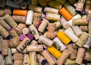 A close-up view of dozens of used wine bottle closures piled together, including natural corks, synthetic corks, and plastic stoppers. The corks vary in color and shape, with many bearing winery names, logos, and vintage markings in black, purple, orange, yellow, and cream tones.