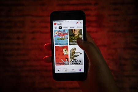 A hand holding a smartphone displaying the YouTube app, with video thumbnails visible on the screen, illuminated by red ambient lighting against a dark, blurred background.