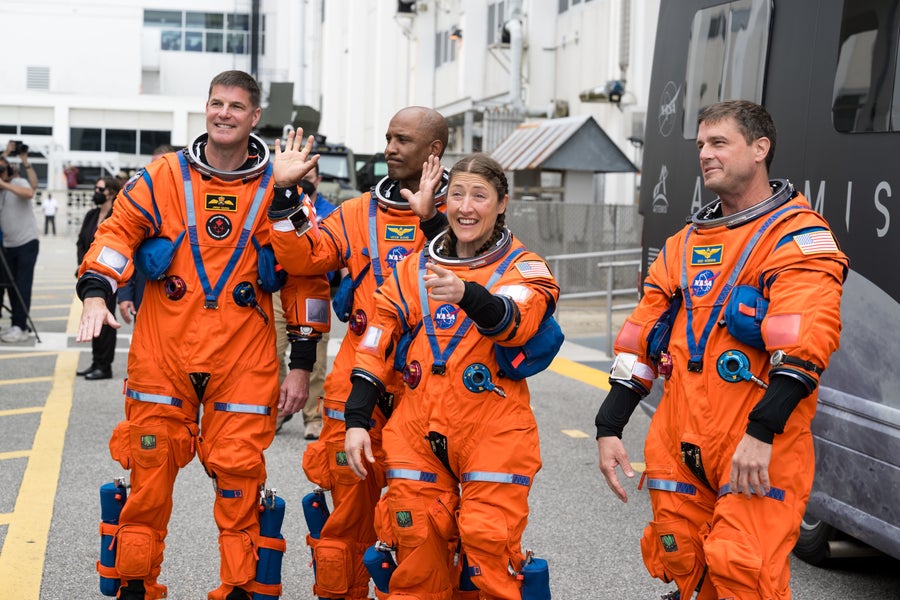 Four astronauts in orange flight suits wave to onlookers.