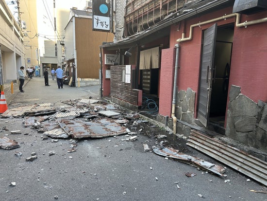 A photograph looking down an alleyway where parts of a wall have collapsed onto the sidewalk. People stand around in the distance.
