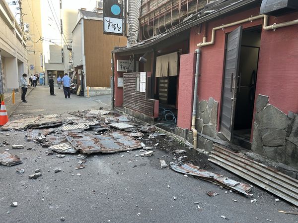 A photograph looking down an alleyway where parts of a wall have collapsed onto the sidewalk. People stand around in the distance.