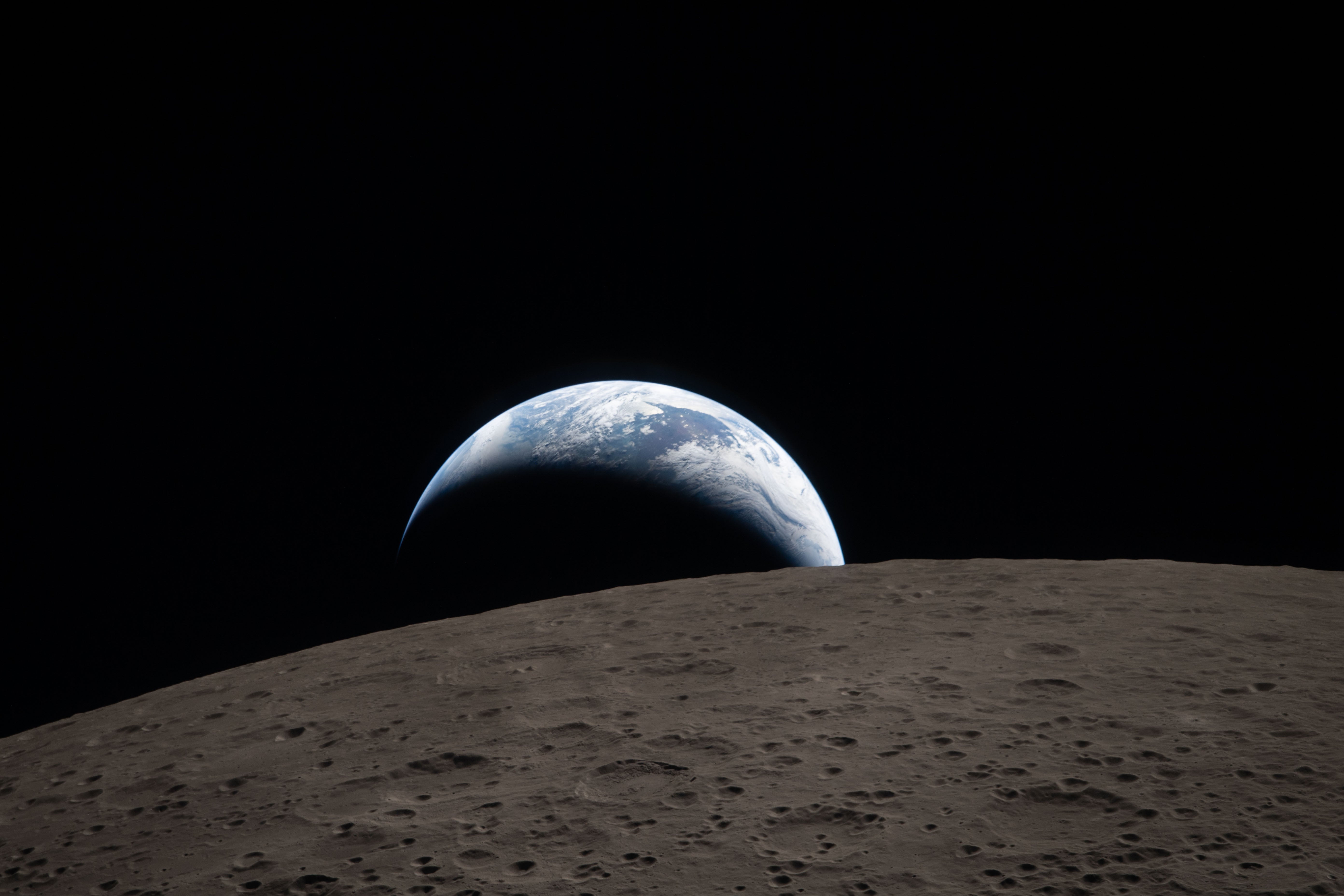 A crescent-lit Earth peeking out over a dark lunar surface like a half-obscured sunrise.