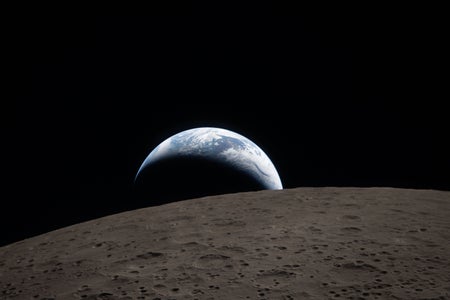 A crescent-lit Earth peeking out over a dark lunar surface like a half-obscured sunrise.