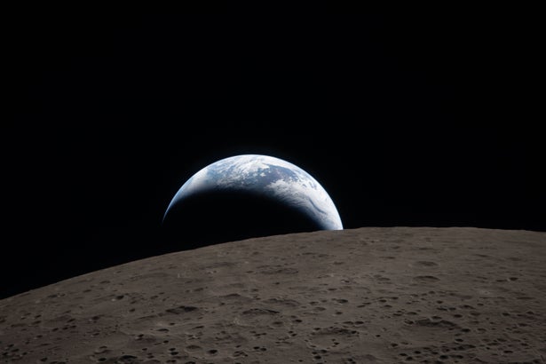 A crescent-lit Earth peeking out over a dark lunar surface like a half-obscured sunrise.