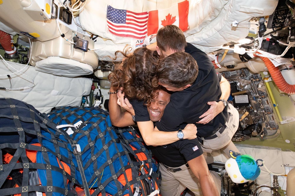 Cuatro astronautas comparten un abrazo grupal dentro de una pequeña nave espacial abarrotada. Un novedoso peluche de la luna con un sombrero terrestre mira con nostalgia.