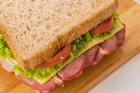 A thick sandwich with ham, cheese, lettuce, and tomato on toasted whole wheat bread, served on a wooden cutting board.